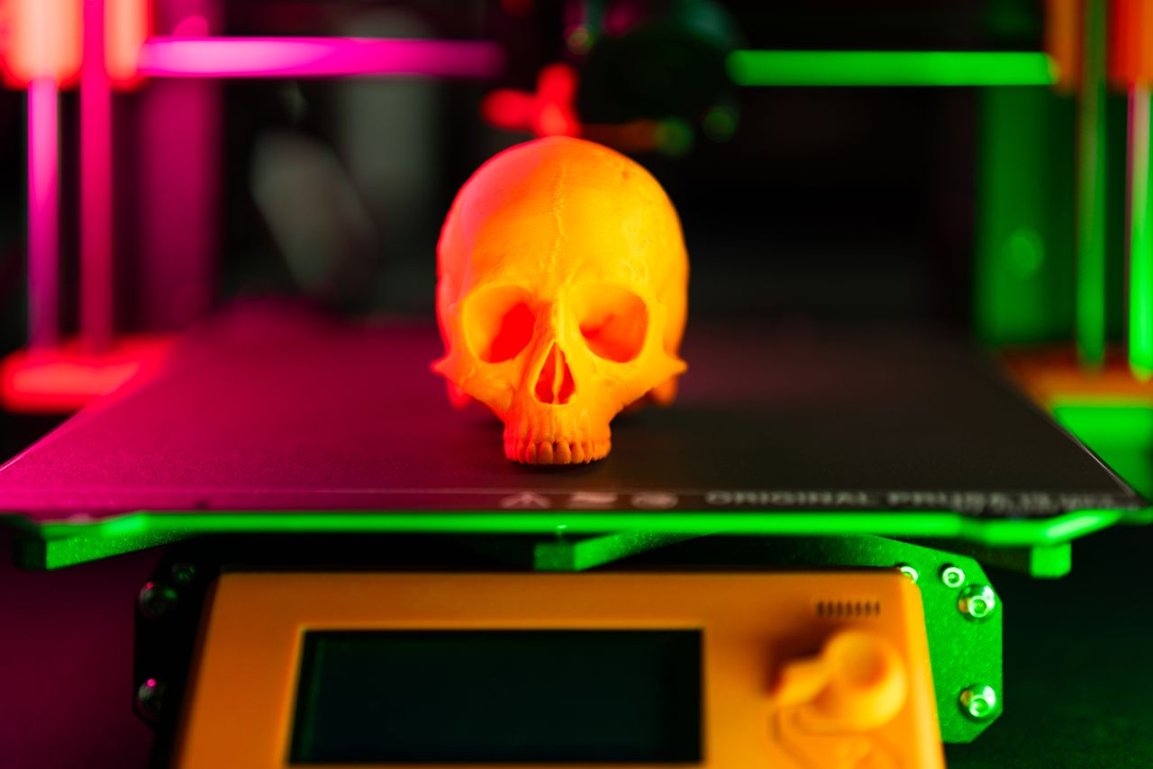 services-01 Close-up of an orange 3D-printed skull on a 3D printer with vibrant lighting.
